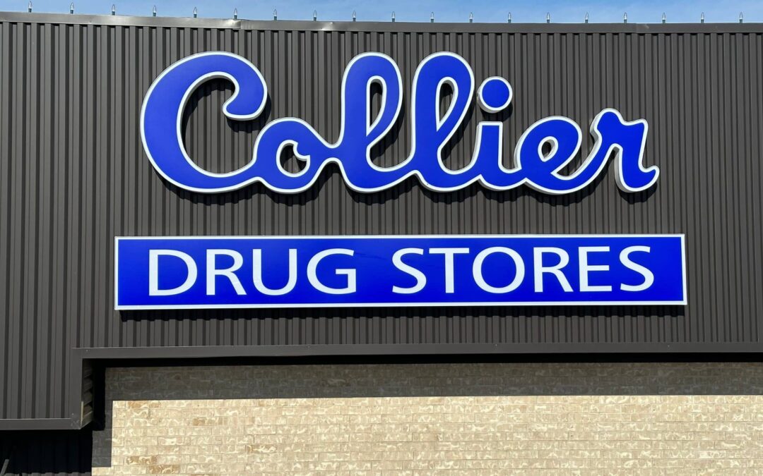 A sign on a building reads Collier Drug Stores in blue and white letters. The text Collier is in a cursive font above DRUG STORES, which is in a bold, block typeface. The background is dark gray metal siding.