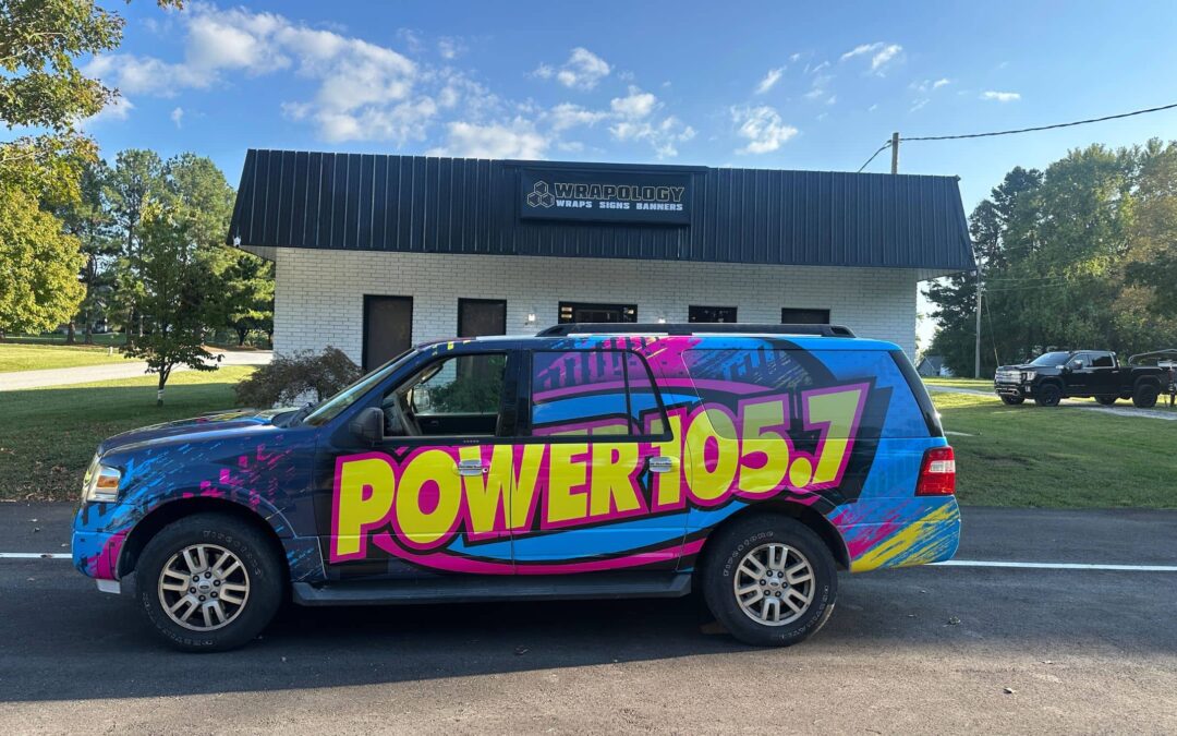 A blue SUV with vibrant pink, yellow, and blue designs featuring POWER 105.7 is parked in front of a building labeled Ampology Wraps Signs Banners under a clear sky. Trees and a few more vehicles are visible in the background.