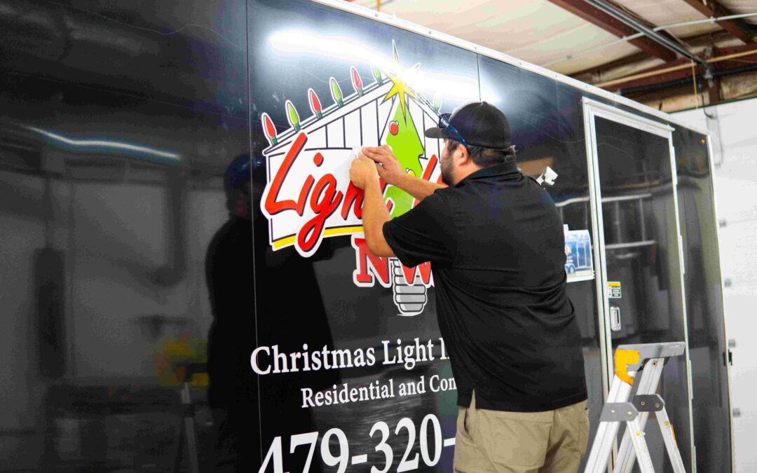A man in a black shirt and cap applies a festive Lights decal to the side of a black trailer advertising Christmas light installation services. A stepladder is nearby and the trailer displays contact information.