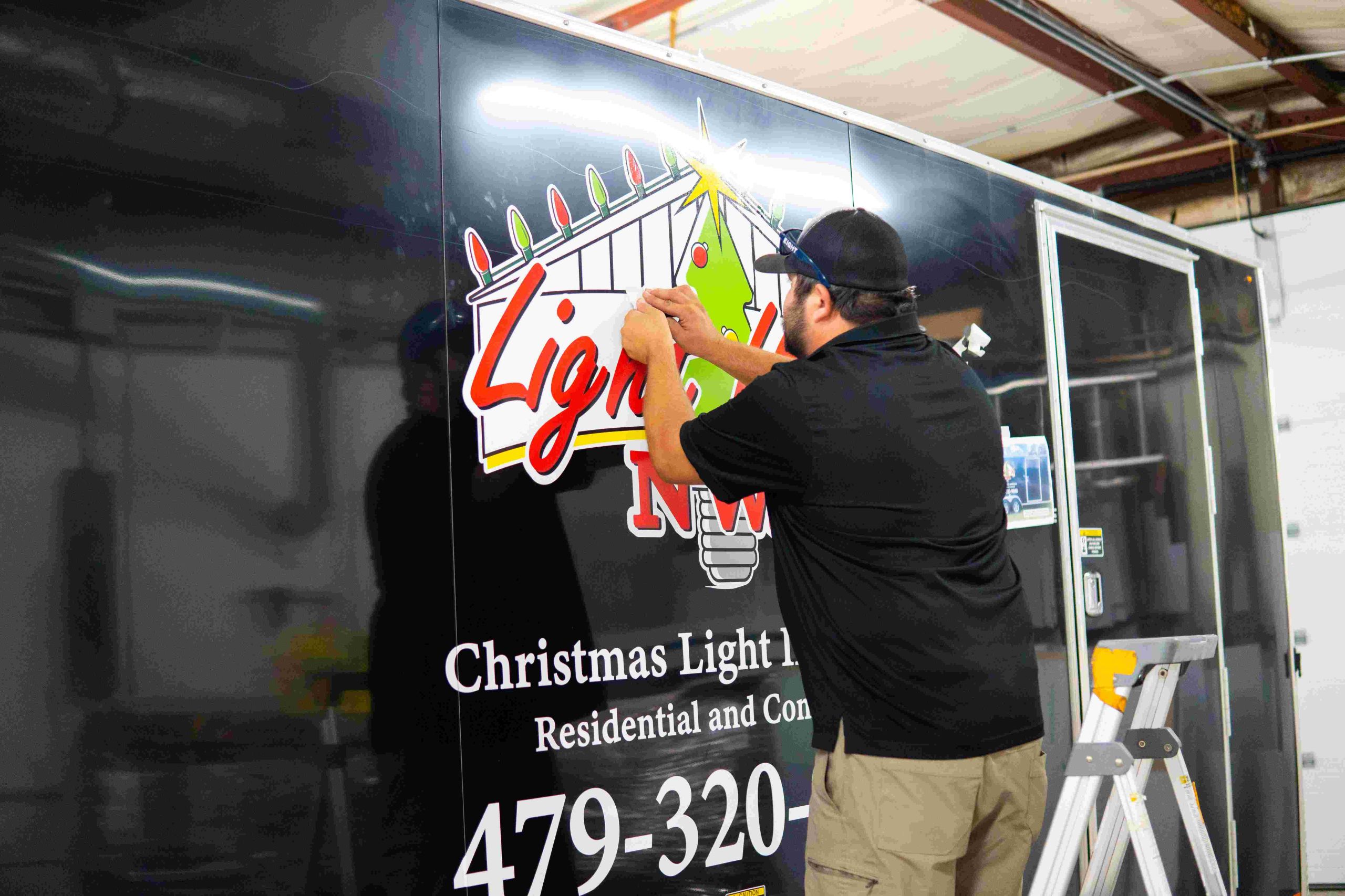 A man in a black shirt and cap applies a festive Lights decal to the side of a black trailer advertising Christmas light installation services. A stepladder is nearby and the trailer displays contact information.