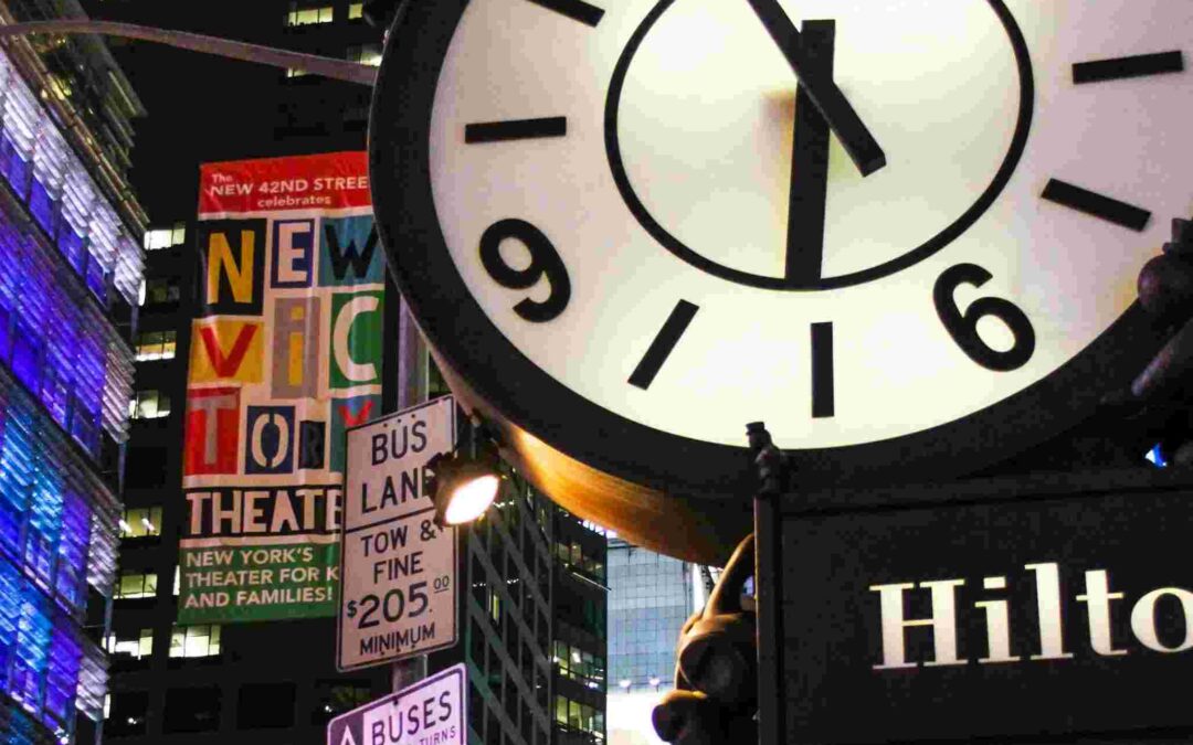 A large clock showing 7:51 is displayed above a Hilton hotel sign in a vibrant city scene at night, with colorful theater advertisements and street signs for buses and parking visible in the background.
