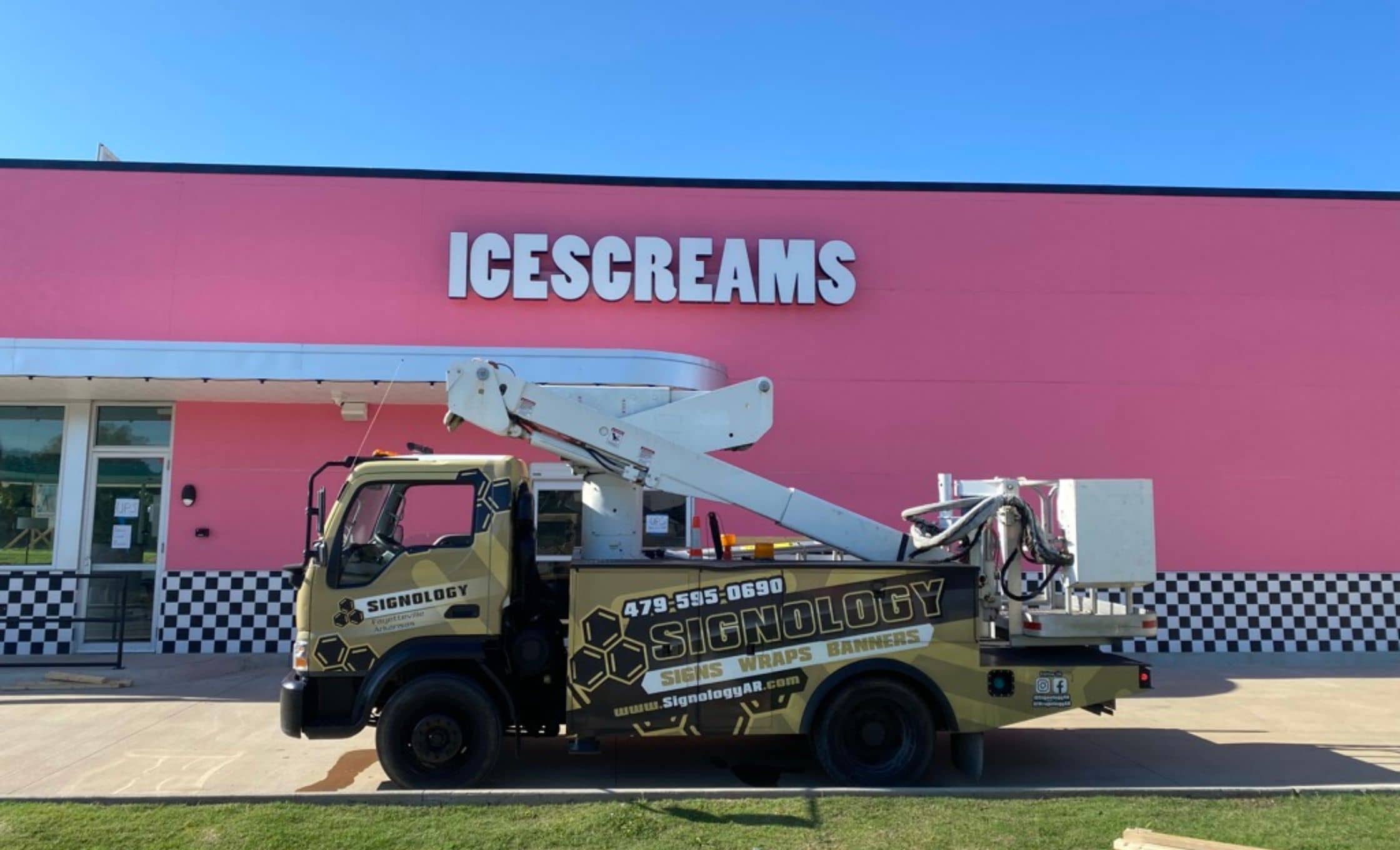 A Signology truck with a mounted lift is parked outside a pink building with a sign that reads ICESCREAMS. The truck displays company info for sign, wrap, and banner services. The sky is clear and blue.