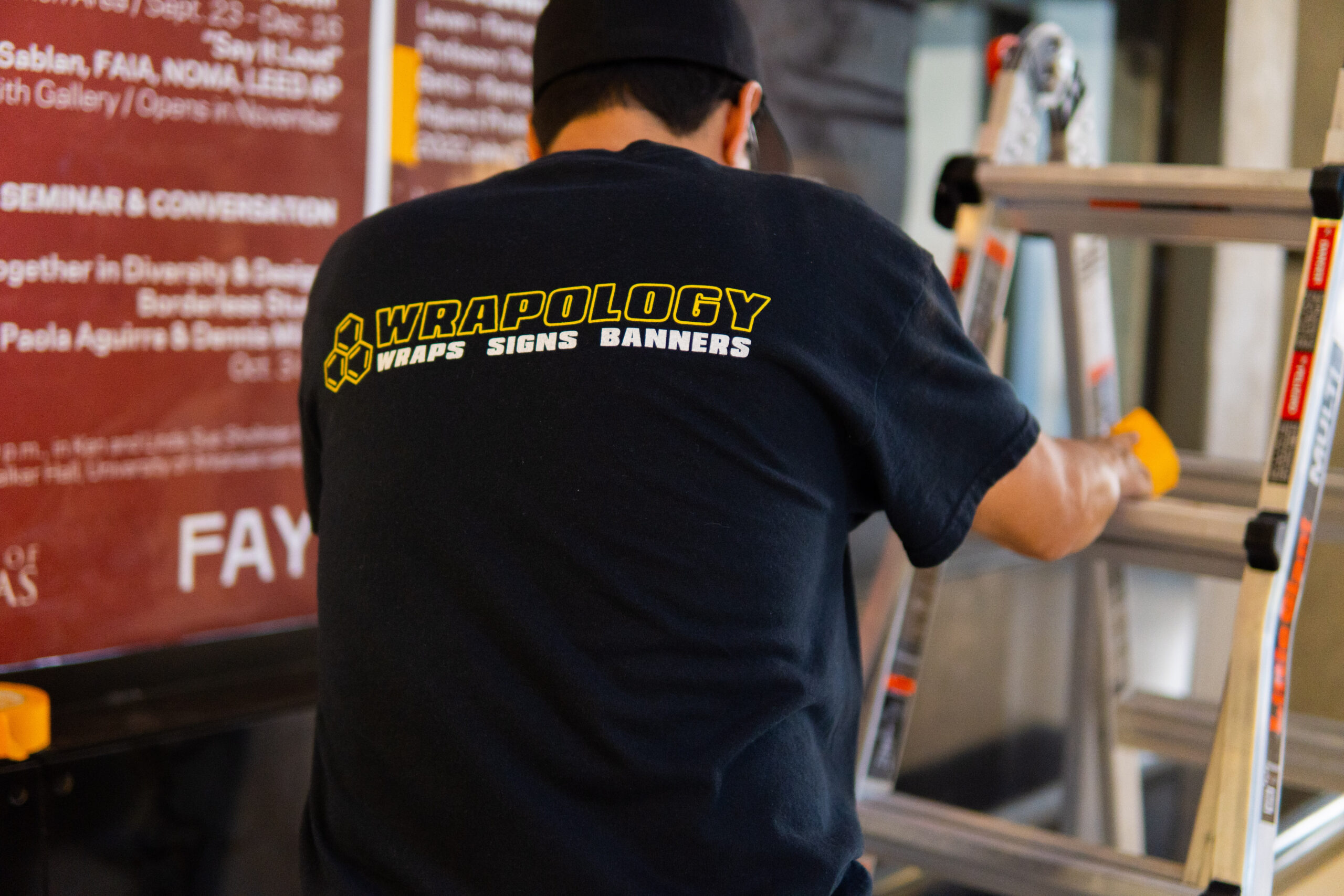 A person wearing a black Wrapology Wraps Signs Banners t-shirt is standing next to a silver ladder, facing away from the camera, with a brown information board visible in the background.