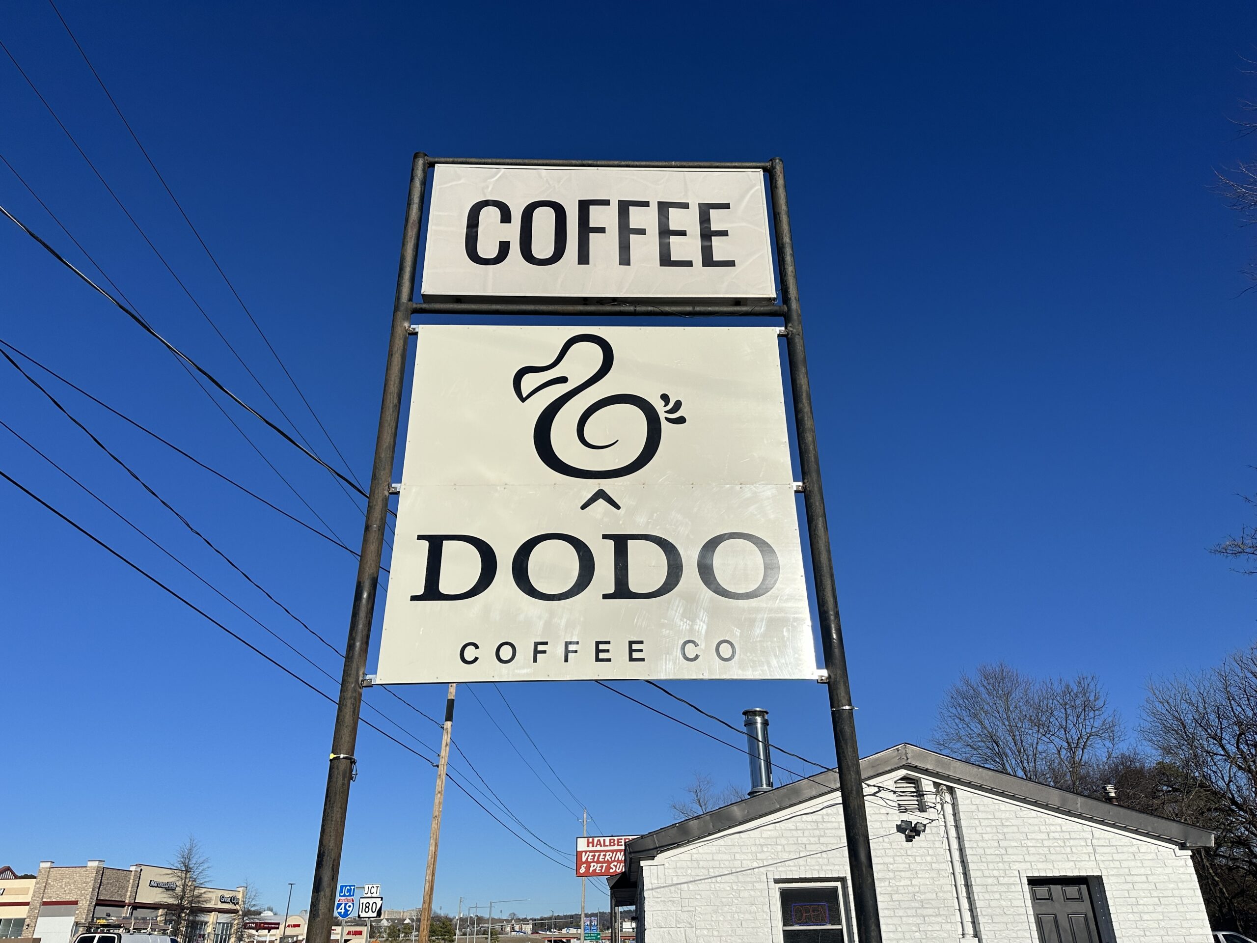 IMG_8509 (1) A sign with a stylized dodo bird logo and the text DODO COFFEE CO sits below a plain COFFEE sign, showcasing creative signage for storefronts under a clear blue sky near a white building.