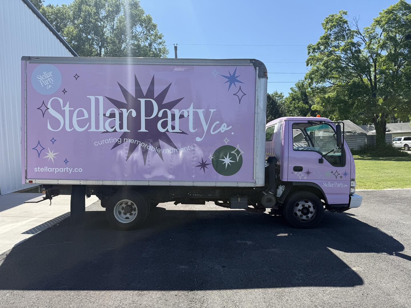 A purple delivery truck with Stellar Party Co. and star designs on the side is parked on asphalt next to a white building, with trees and grass in the background on a sunny day.