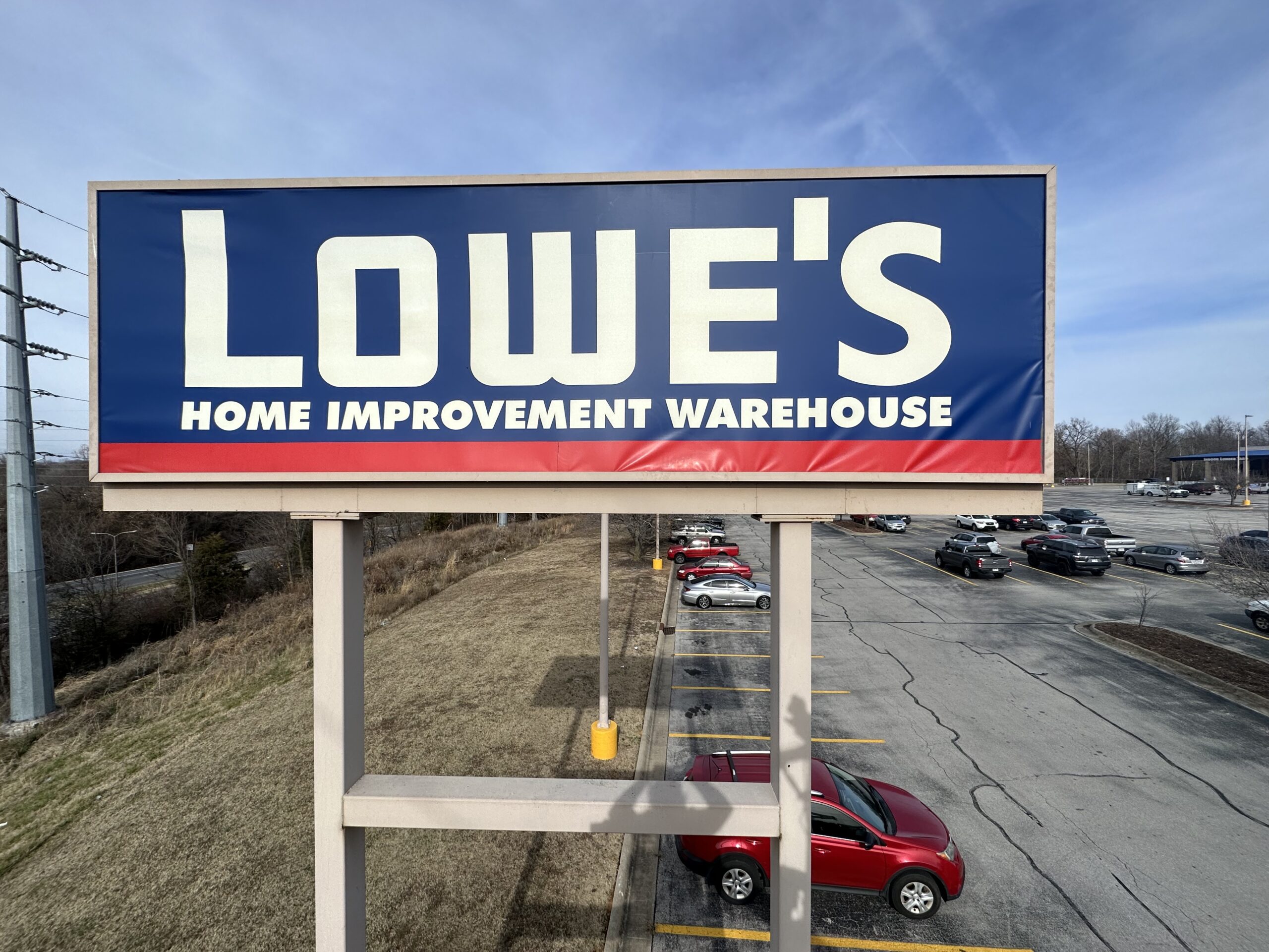 color signage A large outdoor sign for Lowes Home Improvement Warehouse stands over a parking lot with several parked cars, bordered by grass and trees under a blue sky.