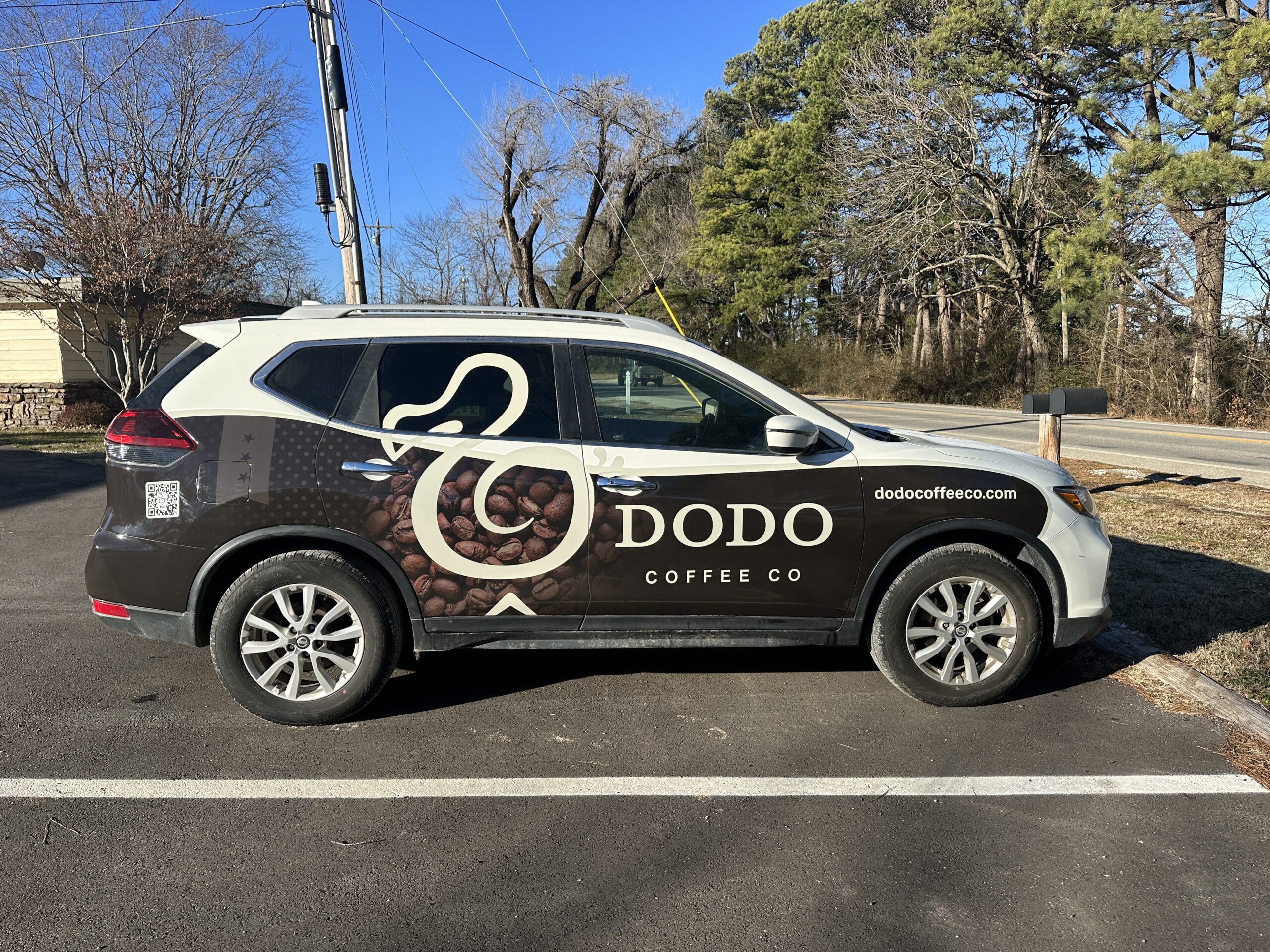 A silver SUV with Dodo Coffee Co branding, coffee bean graphics, and dodocoffeeco.com on its side features weather-resistant wraps, parked in a lot near a road with trees and houses in the background.