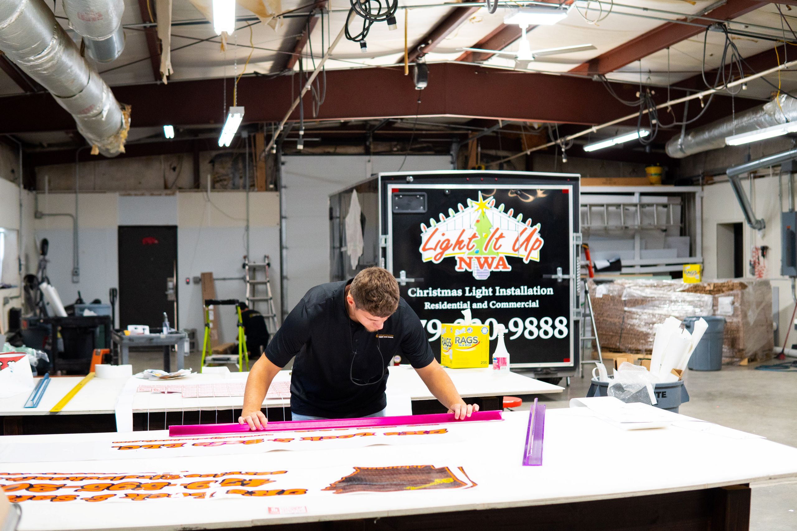 A man works in a warehouse, measuring and aligning decals for custom storefront signs on a table. Behind him is a trailer with the words Light It Up NWA and information about a Christmas light installation business.