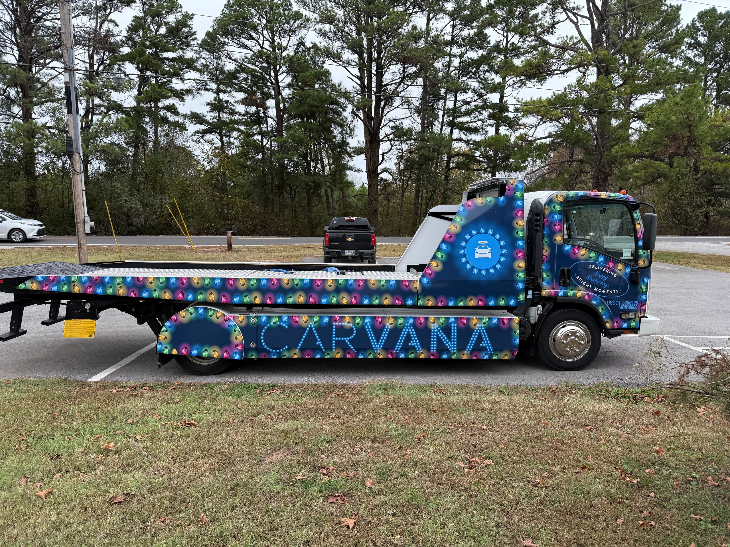 A colorful Carvana flatbed tow truck with vibrant fleet wraps is parked in a lot. The truck features multi-colored circles and the Carvana logo on the door and side. Pine trees and cars are visible in the background.