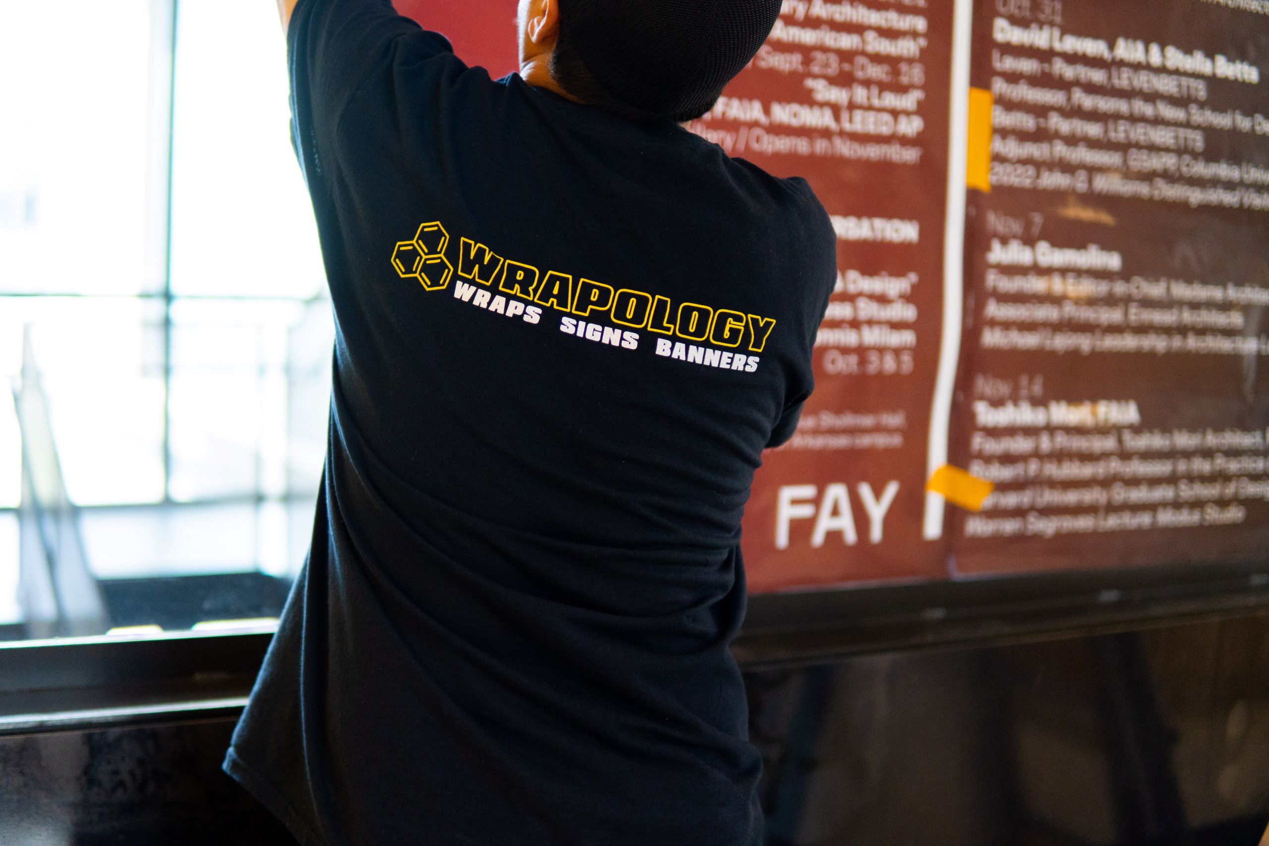 A person wearing a black Wrapology Wraps Signs Banners t-shirt works on installing or adjusting large window graphics against a glass surface. The person's face is not visible.