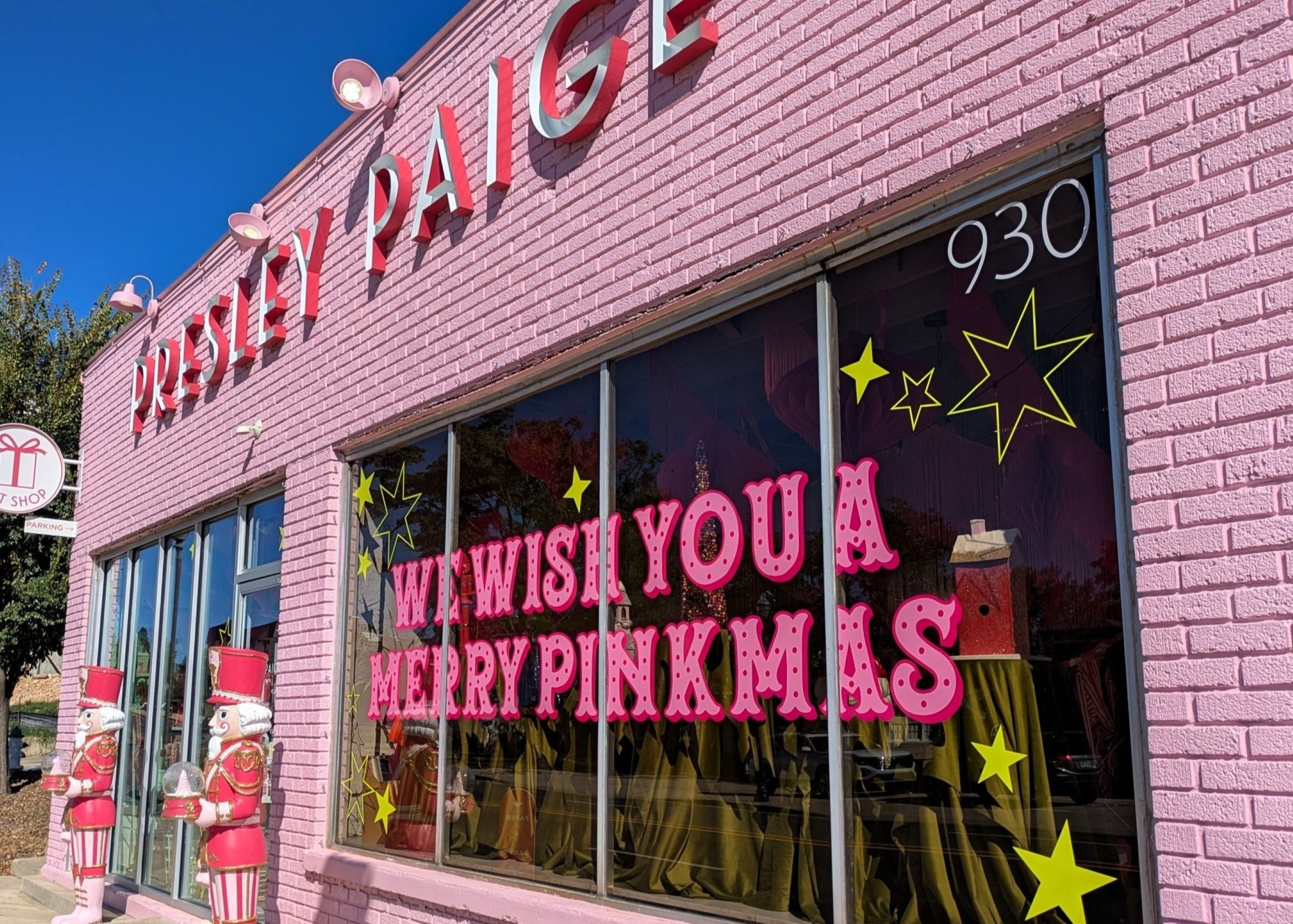 A pink brick storefront with “We wish you a Merry Pinkmas” in large pink letters on the window, yellow stars, nutcracker decorations by the entrance, and hints of year-end rebranding. The store name PINKY PAIGE is above the door.