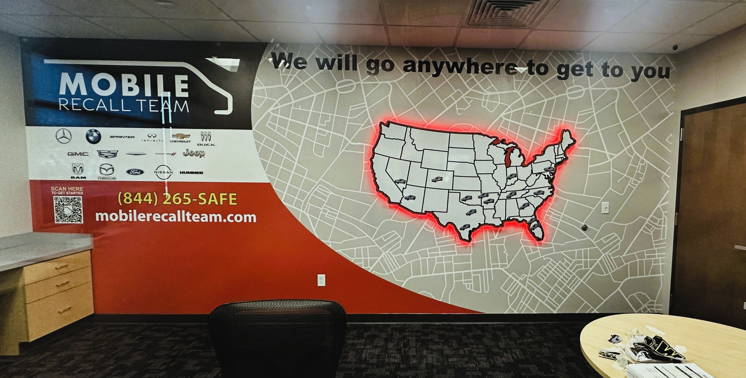 Office wall mural with a U.S. map outlined in red, the words We will go anywhere to get to you, car brand logos, and contact info for the Mobile Recall Team—perfect inspiration for Signage Ideas for 2026 above desks and a chair.