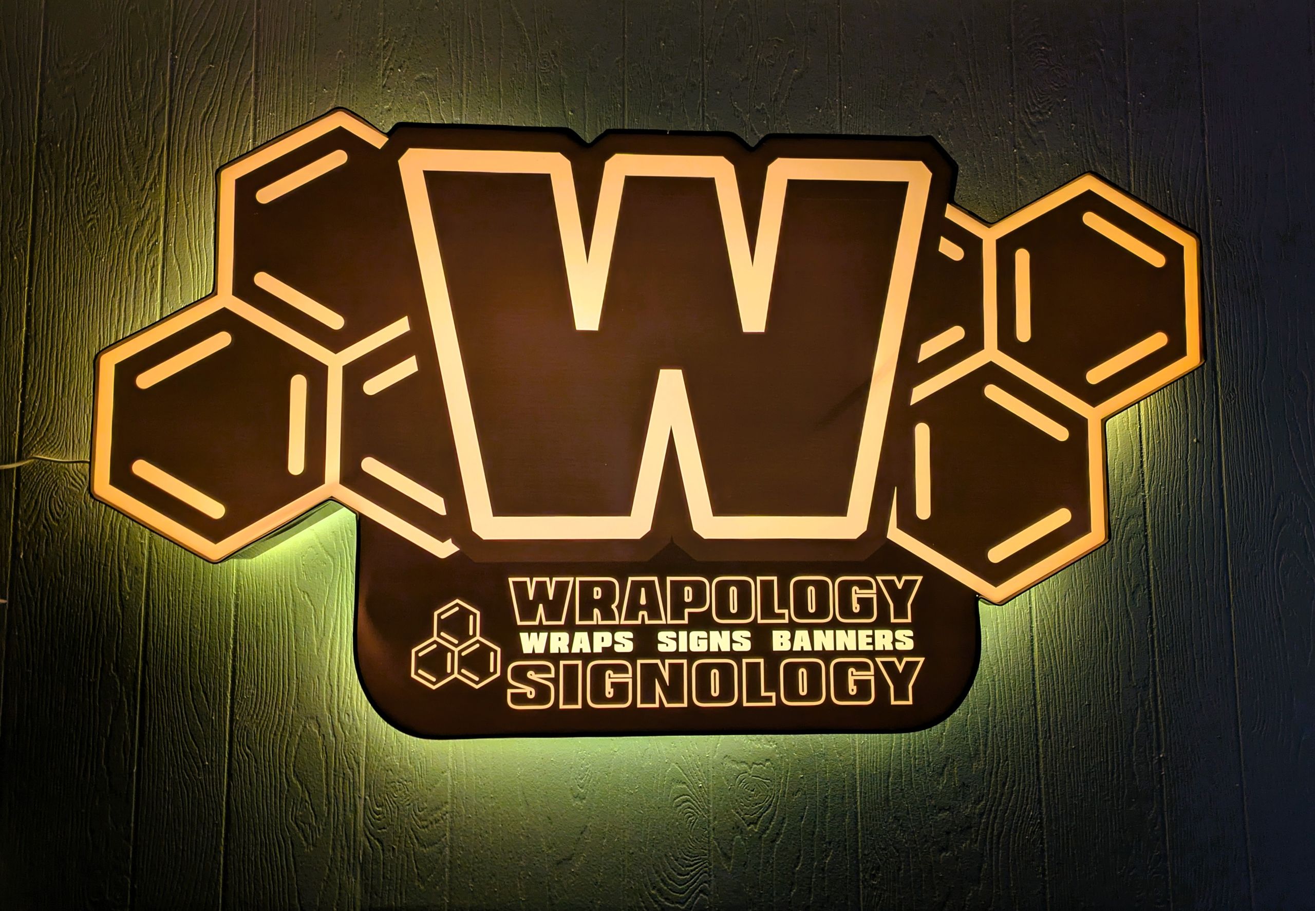 A large illuminated sign displays a bold “W” with honeycomb shapes around it, showcasing quality business signage. Below, text reads: “Wrapology Wraps Signs Banners Signology” in white and yellow letters on a dark background.