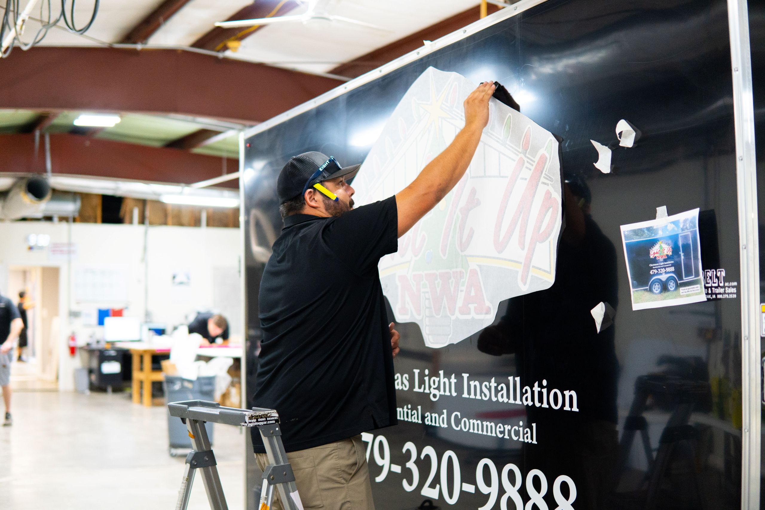 A man wearing a cap and mask applies a large vinyl decal with business information onto a black wall inside a workshop, where tips like "How Long Do Vehicle Wraps Last" might also be shared. A ladder and office area are visible in the background.