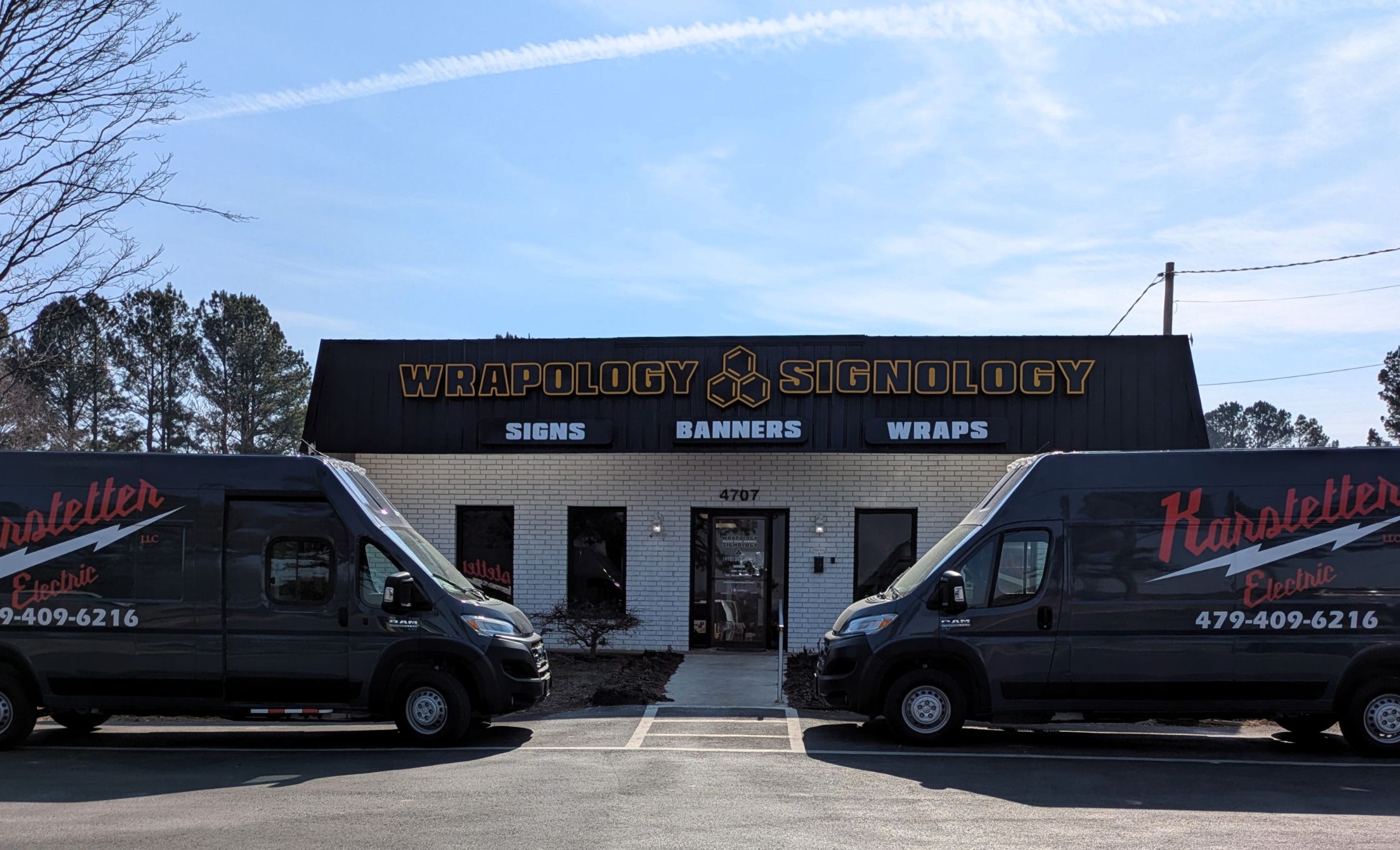 Two black vans with Karstetter Electric parked in front of a brick building with a sign reading Wrapology Signology: Signs, Banners, Wraps. Trees and a blue sky are visible in the background.