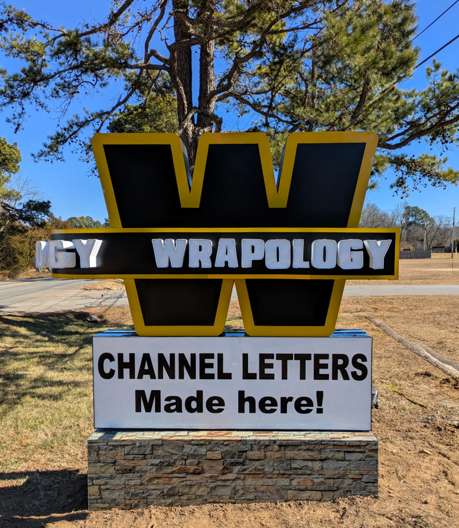 Large outdoor sign with a big yellow and black W and the word WRAPOLOGY. Below, it reads, CHANNEL LETTERS Made here! The sign stands in front of trees and a road.