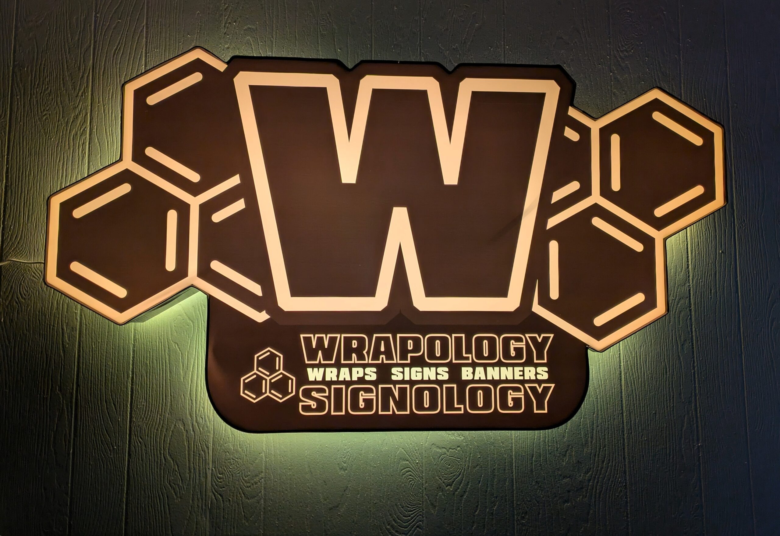 A large illuminated sign with a bold letter W surrounded by hexagonal shapes. Below, it reads WRAPOLOGY Wraps Signs Banners SIGNOLOGY in capital letters. The sign is mounted on a dark textured wall.