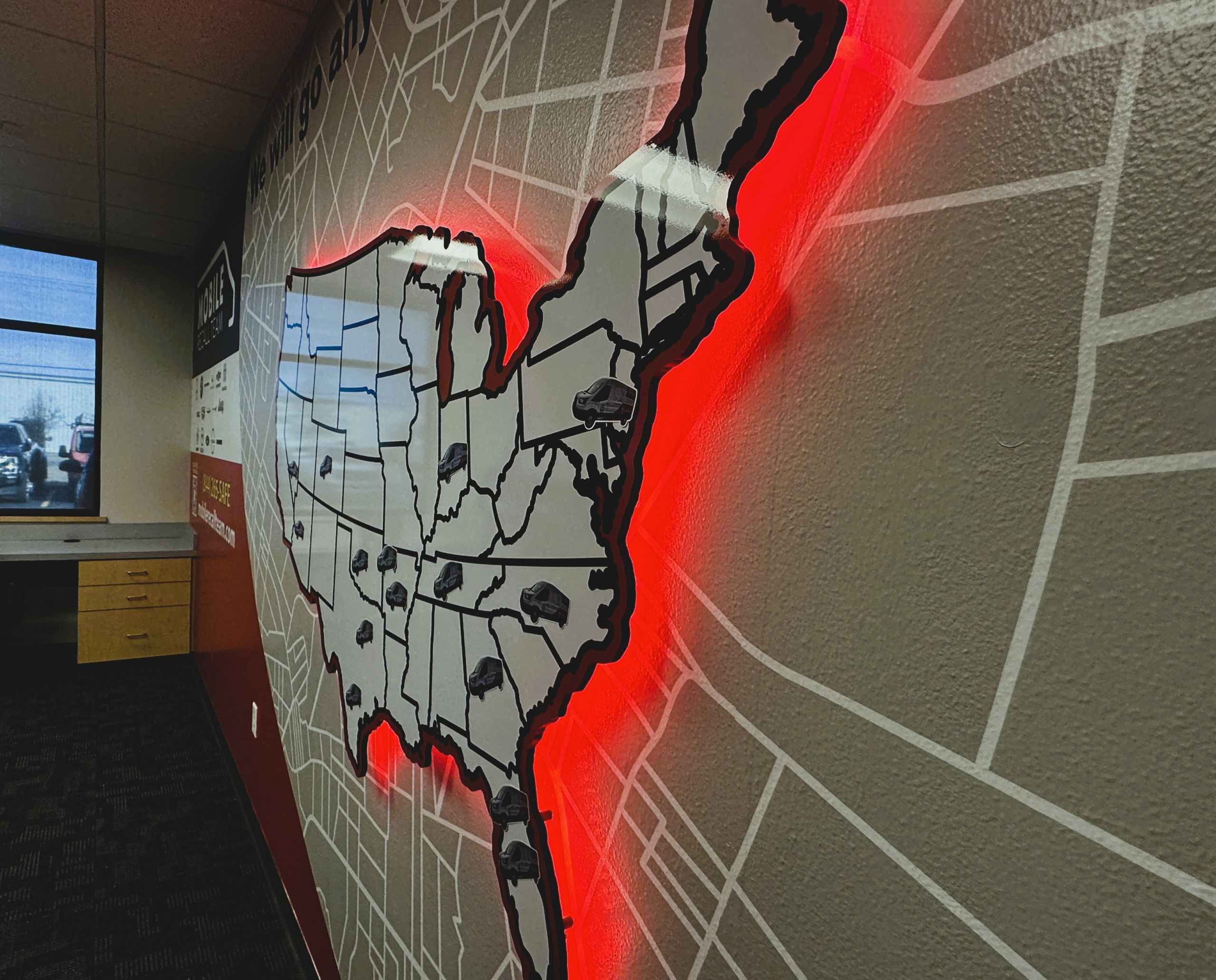 A wall map of the United States outlined with a bright red light, featuring small car icons marking various cities. This striking piece of indoor branding is displayed in an office, with desks and windows visible in the background.