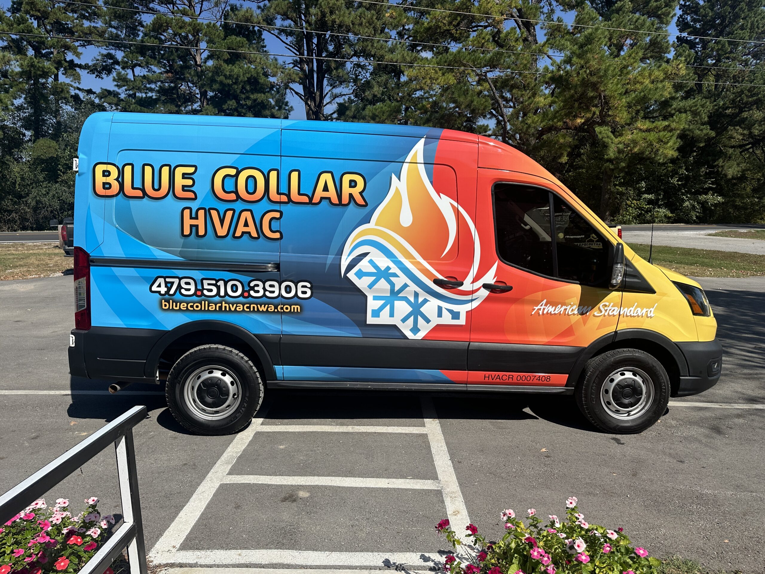 A colorful van with a vibrant vehicle wrap for Blue Collar HVAC and contact info on the side, featuring a large flame and snowflake graphic, parked in a lot with trees in the background.