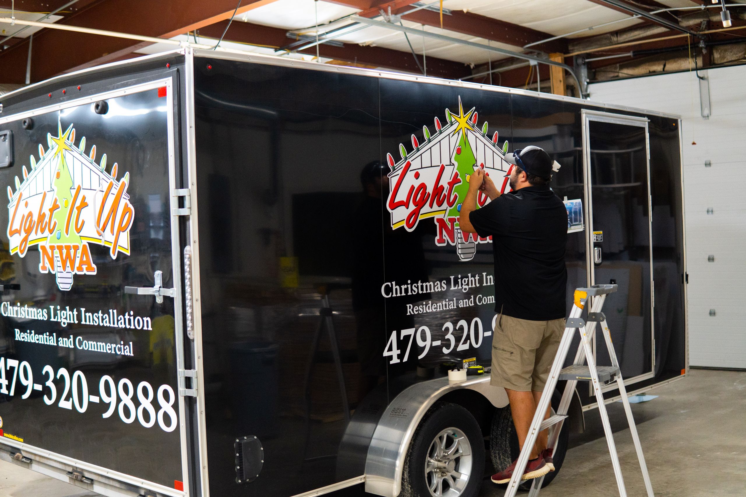 A man stands on a step ladder, applying decals to a black trailer with Light It Up NWA: Christmas Light Installation, Residential and Commercial printed on the side—showing how you can wrap your vehicle for professional branding inside a large garage.