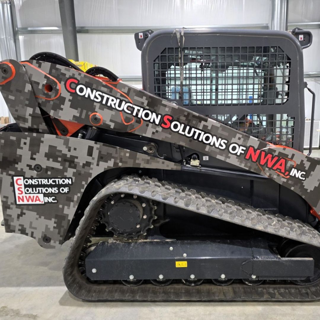 Construction Solutions - Square A skid steer loader with a digital camouflage wrap and “Construction Solutions of NWA, Inc.” written on the side, parked indoors on a concrete floor.