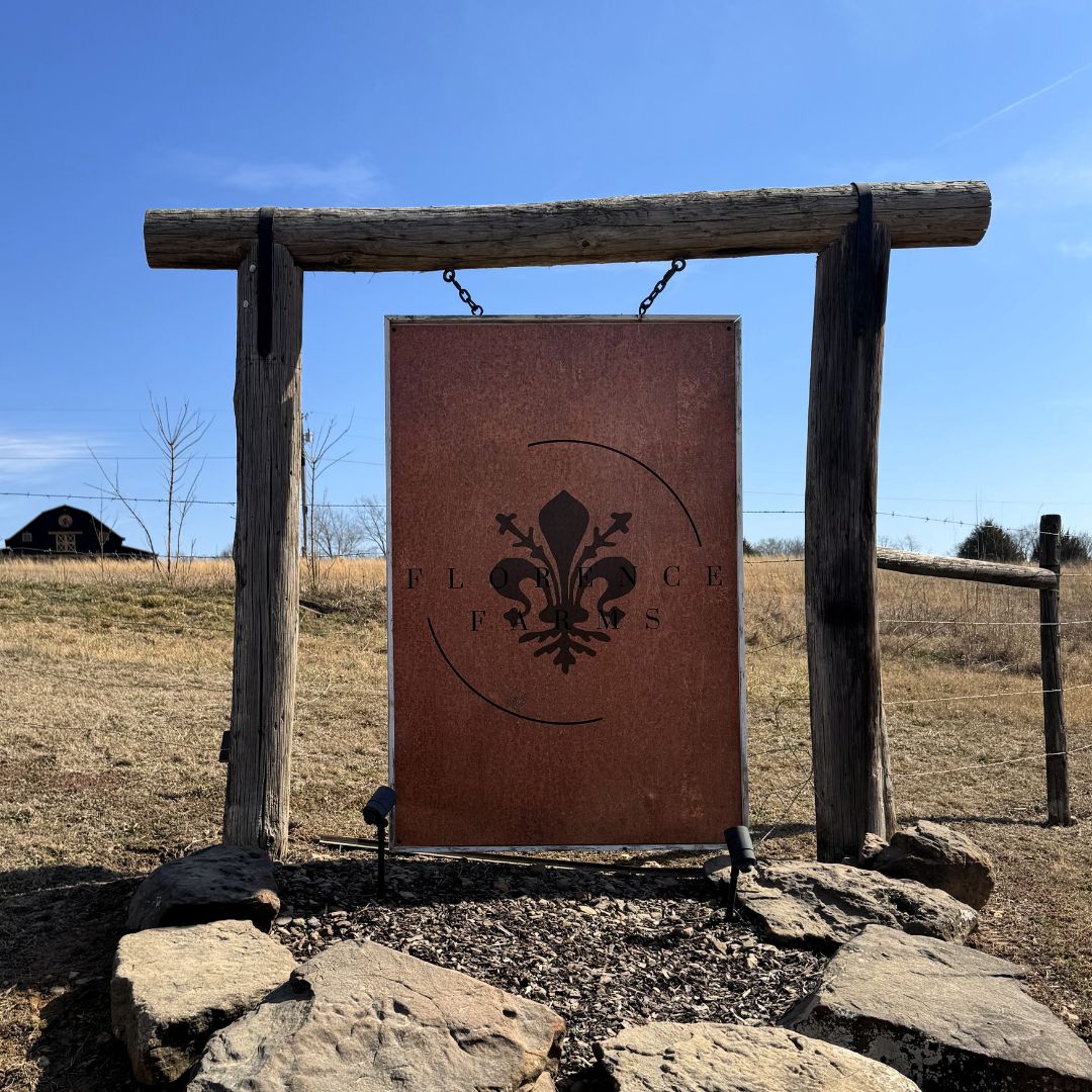 A rustic wooden sign with a fleur-de-lis design and the words FLORENCE F.S. stands on a grassy, open field under a clear blue sky. There is a barn in the background to the left.