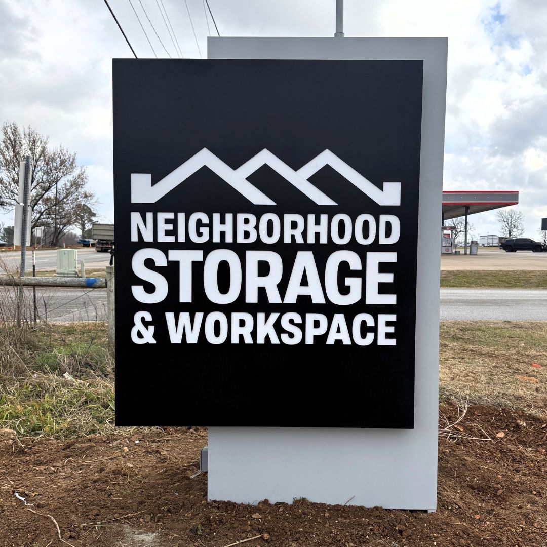 A black and white outdoor sign reads Neighborhood Storage & Workspace with a stylized roof design above the text, located near a road with grass and some trees in the background.
