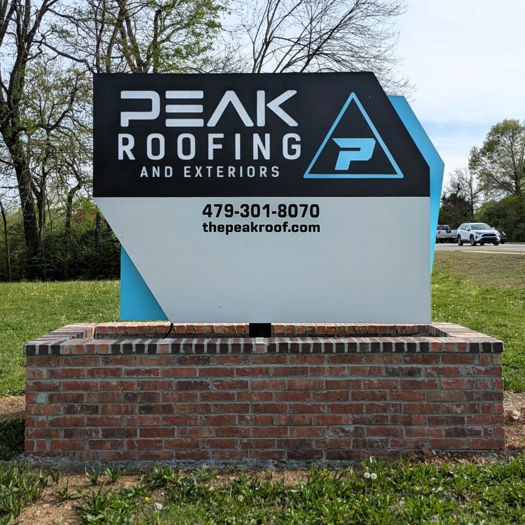 A sign for Peak Roofing and Exteriors sits on a brick base, displaying their logo, phone number (479-301-8070), and website (thepeakroof.com), with trees and cars visible in the background.