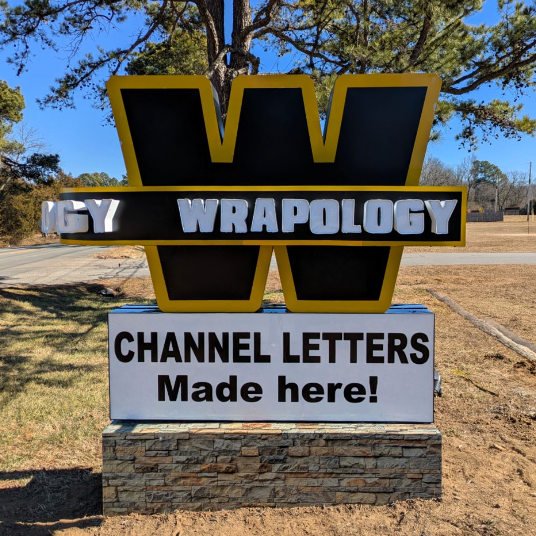 A large sign with a bold yellow and black W and the word WRAPOLOGY in silver letters. Below, a white panel reads: CHANNEL LETTERS Made here! The sign is outdoors on a stone base near trees and grass.