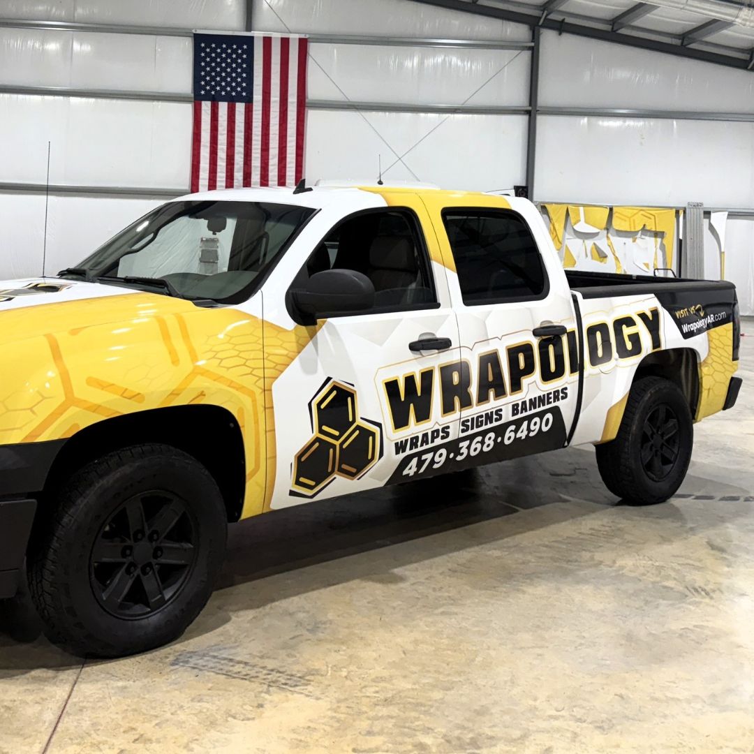 Wrapology Truck - Square A white pickup truck with a yellow and black honeycomb-themed wrap displaying the business name Wrapology and contact number 479-368-6490, parked inside a garage with an American flag on the wall.