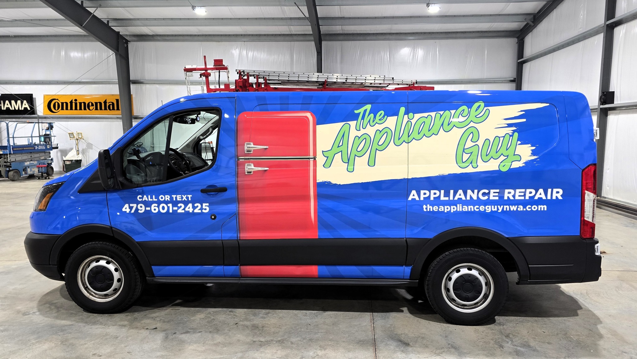 A blue van with The Appliance Guy logo, a red refrigerator graphic, and contact info for appliance repair services, parked inside a garage. The phone number and website are visible on the side of the van.
