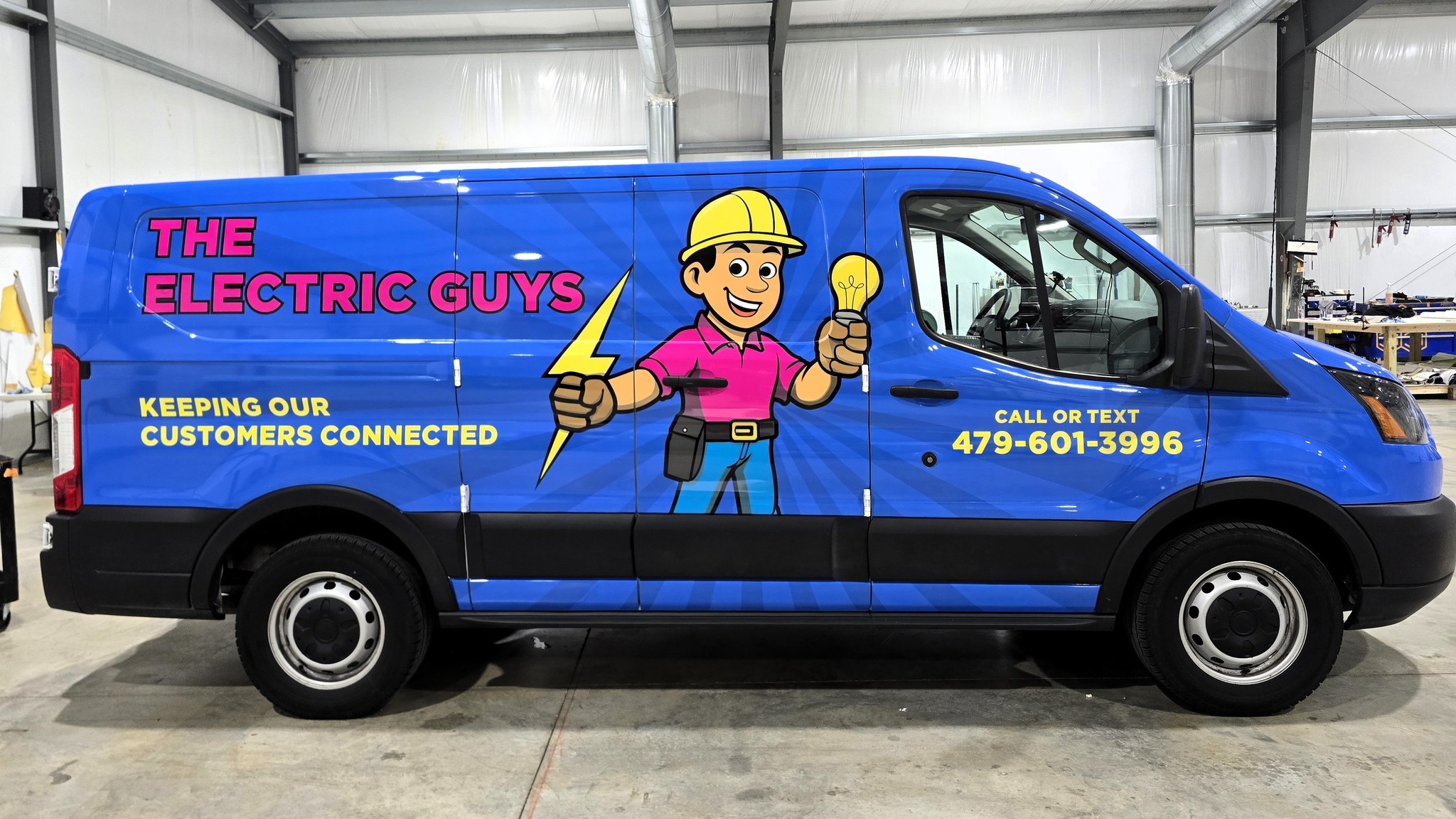 A blue van with “The Electric Guys” in bold pink and white letters, a cartoon electrician holding a lightbulb and lightning bolt, and contact info: “Call or text 479-601-3996.” Slogan reads “Keeping our customers connected.”.