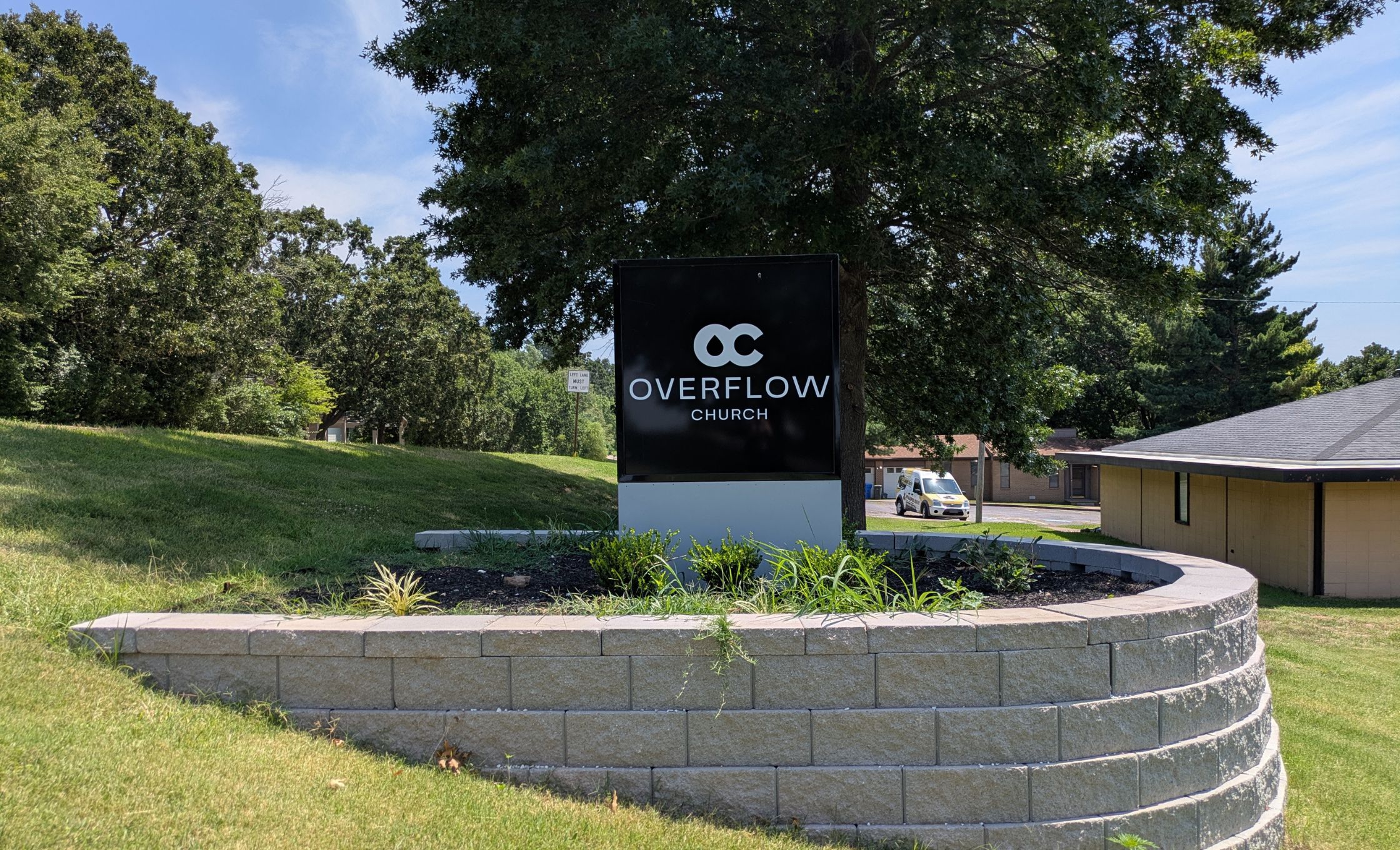 A sign reads Overflow Church with a logo above it, set on a landscaped stone platform with grass, plants, and trees in the background.