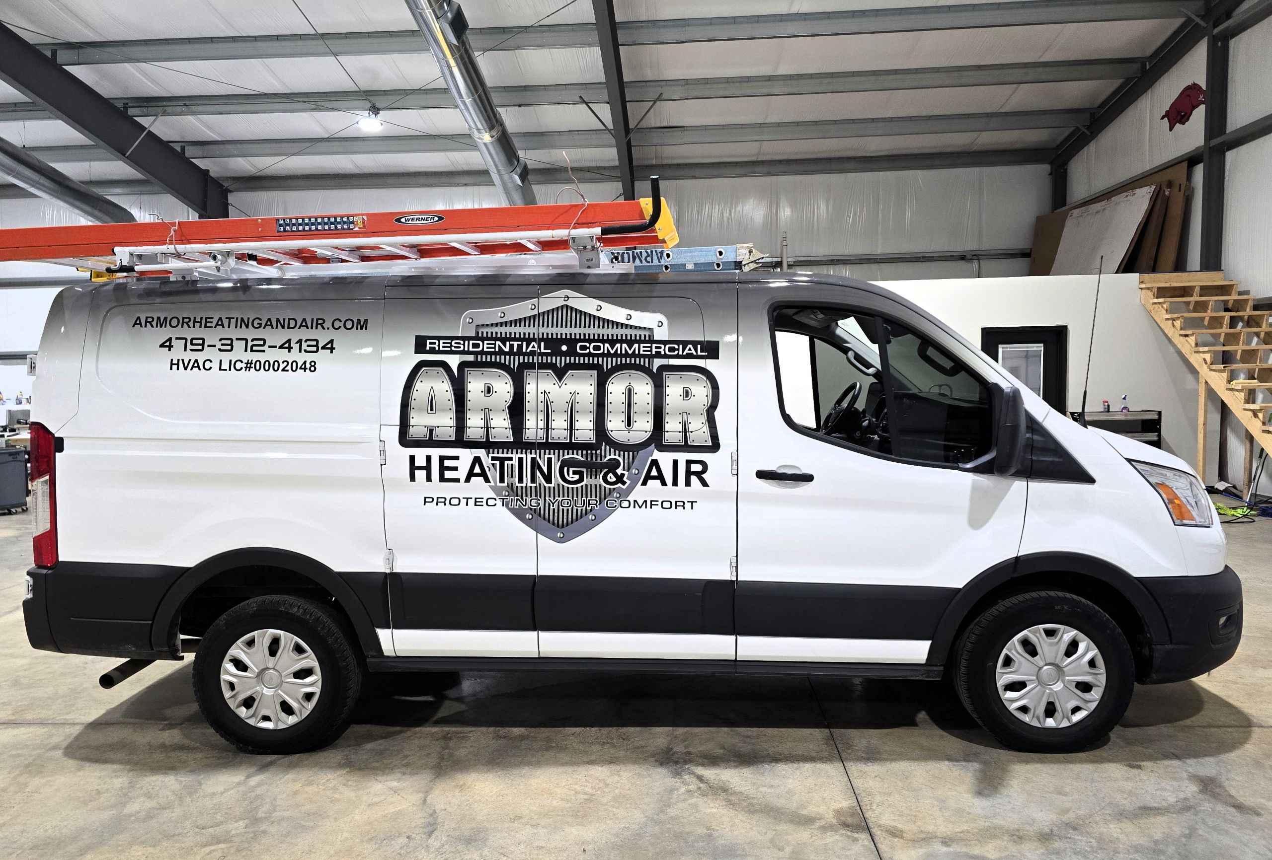How Commercial Vehicle Wrapping Helps Businesses Stay Competitive White van with Armor Heating & Air branding, a roof-mounted orange ladder, and business contact details on the side, parked inside a large industrial garage with metal beams and a wooden staircase.