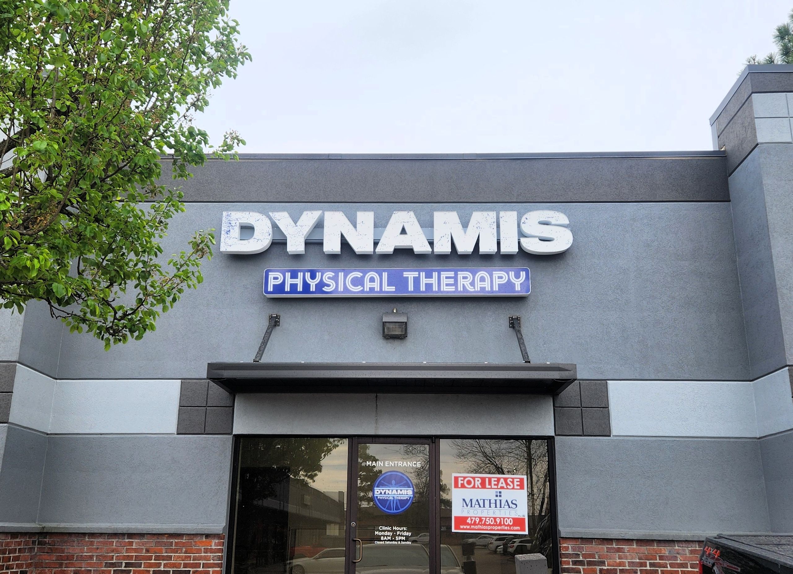 The Benefits of Door Graphics for Retail Stores and Office Spaces Storefront of Dynamis Physical Therapy with large signs above the entrance. A For Lease sign is visible on the entry door. A tree is on the left side of the image. The building exterior is gray with brick accents.
