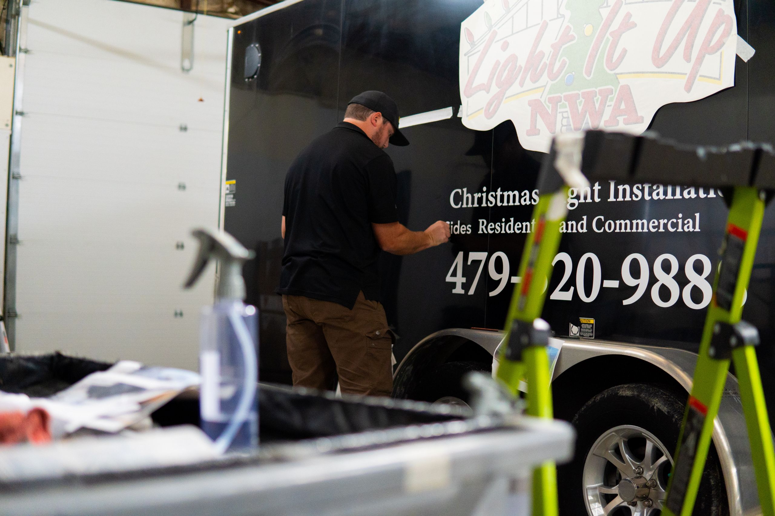 Vehicle Wraps in Fayetteville What Local Businesses Should Know A man in a black shirt and cap works on a black trailer labeled Light It Up NWA Christmas Light Installation inside a garage. Cleaning supplies and a green ladder are in the foreground.