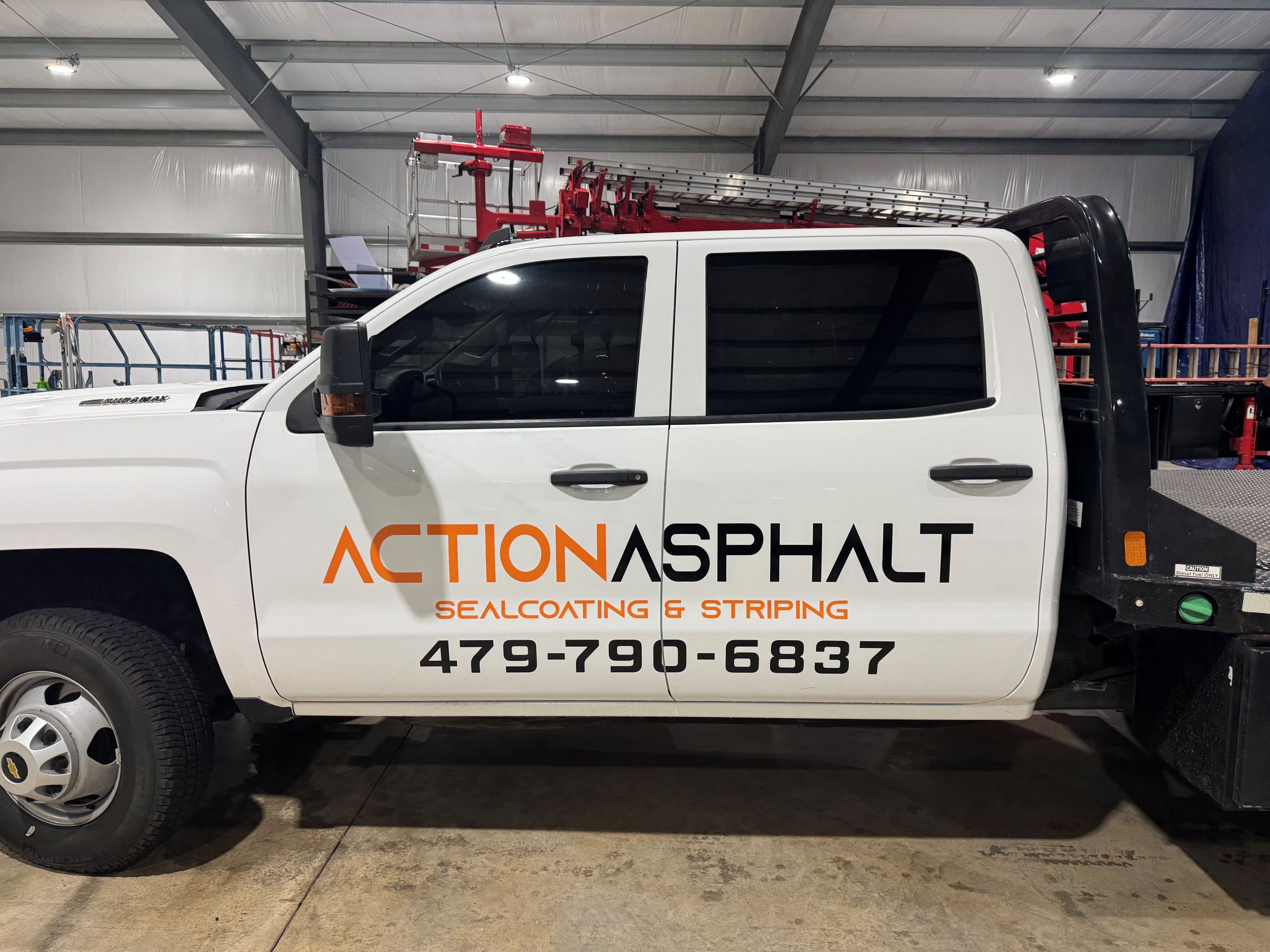 What Happens If You Wait Too Long to Wrap Your Work Truck White truck with Action Asphalt Sealcoating & Striping and phone number 479-790-6837 on the door. Red ladders are stacked on a rack in the truck bed. The truck is parked inside a large garage.
